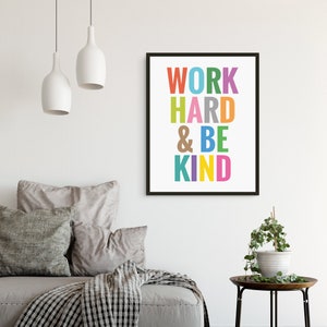 May include: A framed print with the text "WORK HARD & BE KIND" in colorful letters on a white background. The print is hanging on a white wall above a gray couch with pillows.
