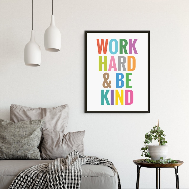 May include: A framed print with the text "WORK HARD & BE KIND" in colorful letters on a white background. The print is hanging on a white wall above a gray couch with pillows.