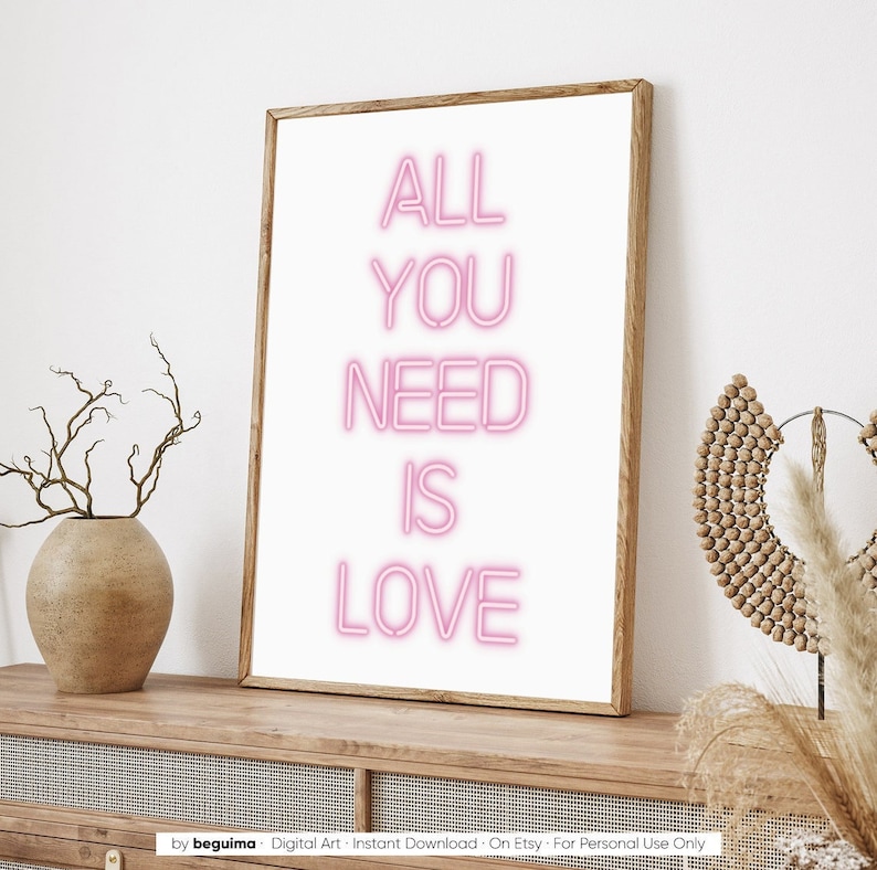 May include: A framed art print with the words "ALL YOU NEED IS LOVE" in pink neon-style lettering on a white background. The wooden frame sits on a wooden cabinet, with a vase and decorative items on either side.
