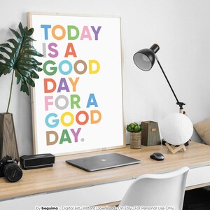May include: A framed print with a white background and colorful text that reads "TODAY IS A GOOD DAY FOR A GOOD DAY." The print is on a desk with a laptop, a lamp, and a potted plant.