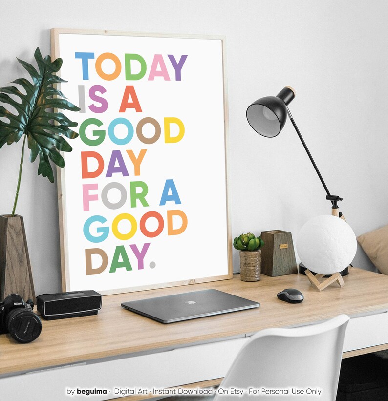 May include: A framed print with a white background and colorful text that reads "TODAY IS A GOOD DAY FOR A GOOD DAY." The print is on a desk with a laptop, a lamp, and a potted plant.