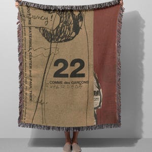 Peut inclure: Une couverture beige et bordeaux avec une bordure à franges. La couverture présente un grand numéro 22 noir, le texte "COMME des GARCONS SUPERDADA" et une illustration de type esquisse. Le texte "BOUT THE WATERMILL CENTER and PAUL THEK" est également visible.