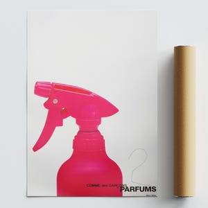 Peut inclure: Une bouteille de pulvérisation rose vif avec une gâchette, sur fond blanc. L'affiche porte le texte "Comme des Garcons Parfums". Un poster enroulé est à droite.