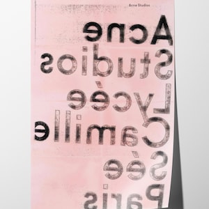 May include: A pink poster with the text "Acne Studios" at the top. The poster features the words "Acne Studios Lycée Camille See Paris 2169" in a distressed, reversed font. The poster has a textured, faded appearance.