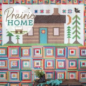 May include: A quilt with a log cabin design and the words "Prairie Home" in a circle. The quilt is made up of squares of different colors and patterns, including stars, stripes, and polka dots. The quilt is displayed on a wooden chest with a smaller quilt on top of it.