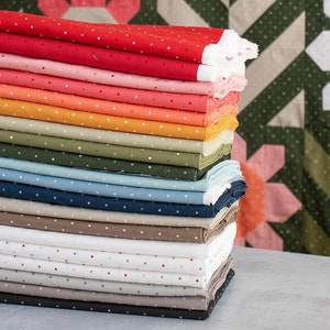 May include: A stack of 15 fabric squares with white polka dots in various colors, including red, orange, pink, green, blue, and brown. The fabric squares are stacked on a light gray surface in front of a colorful quilt.