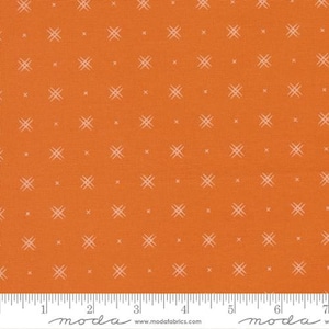 May include: Orange fabric with a repeating pattern of small, light-colored cross-like shapes and dots. The fabric has a textured appearance and a ruler is visible along the bottom edge, marked in inches.