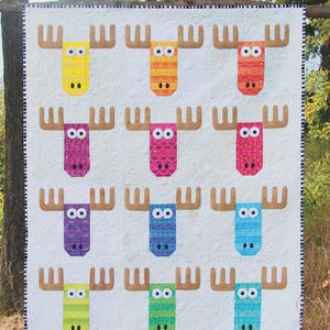 May include: A colorful quilt featuring a repeating pattern of moose with large antlers. Each moose is a different color, including yellow, orange, red, purple, pink, blue, and green. The quilt is labeled "Wild Moose Chase" and "A Quilt Pattern by: Sassafras Lane Designs."