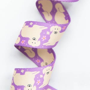 May include: A purple ribbon with a repeating pattern of cream-colored dog silhouettes and small pink flowers. The ribbon is curled, showing the design on both sides. The ribbon is likely used for crafting or gift wrapping.