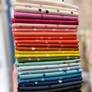 May include: A stack of folded fabric squares in various colors, each adorned with star patterns. The color palette ranges from light pink to deep navy and black. The fabric is likely cotton or a similar material, suitable for quilting or crafting projects.