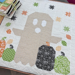 May include: A white quilt featuring a central beige ghost figure with a small geometric pattern. The design includes orange, green, and black patterned pumpkins, along with scattered star shapes in various colors. The fabric has a quilted texture.