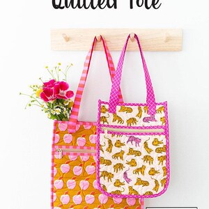 May include: Two quilted tote bags hanging on a wooden rack. One bag is pink with a yellow apple print and the other is pink with a white tiger print. Both bags have pink and white striped straps and gold zippers. The text "Knot + Thread Design" is visible in the bottom right corner.