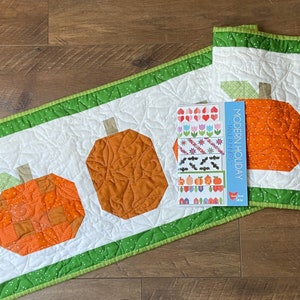 May include: A white quilted table runner with a green border and three orange pumpkin designs. The runner is accompanied by a printed pattern booklet titled "Modern Holiday - Table Runners" with various pumpkin, heart, flower, and bat designs.