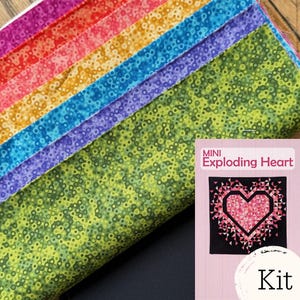 May include: A stack of folded fabric strips in a rainbow gradient of red, orange, yellow, blue, purple, and green, each featuring a small circular pattern. Next to the fabric is a rectangular card labeled MINI Exploding Heart Kit.