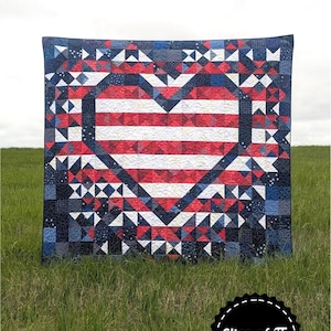 May include: A patriotic quilt with a heart-shaped design, featuring red and white stripes in the center and a blue border with red and white accents. The quilt is displayed on a grassy field. Text on the quilt says "Patriotic Exploding Heart".
