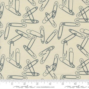 May include: A beige fabric with a repeating pattern of blue safety pins. The fabric has a ruler with the words "moda" and "www.modafabrics.com" printed on it.