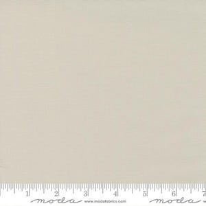May include: A solid, light beige fabric with a subtle woven texture. The fabric is displayed with a ruler along the bottom edge, marked in inches. The fabric is from Moda Fabrics.