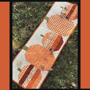 May include: A quilted table runner with a pumpkin theme. The runner features orange pumpkins with green stems and various patterned sections, including gingham and floral designs, set against a cream background. The text "Pumpkin Hollow" is at the top.