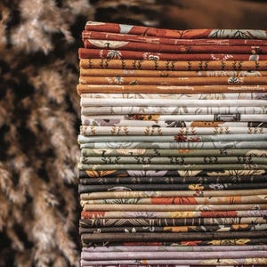 May include: A tall, vertical stack of folded fabric squares featuring a variety of patterns including botanical motifs, pumpkins, and geometric shapes in a color palette of rust, mustard, sage green, cream, brown, plum, and deep charcoal gray.