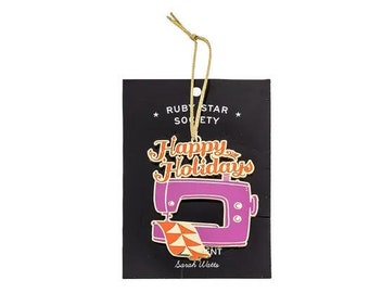 May include: A pink and gold metal ornament shaped like a sewing machine with the text "Happy Holidays" on top. The ornament is from Ruby Star Society and designed by Sarah Watts.