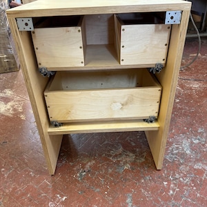 K&ouml;nnte beinhalten: Ein Holzschrank mit zwei Schubladen und zwei offenen F&auml;chern. Der Schrank ist aus hellem Holz gefertigt und hat Metallhalterungen zur Verst&auml;rkung. Die Schubladen sind rechteckig und haben ein schlichtes Design. Der Schrank dient wahrscheinlich zur Aufbewahrung.