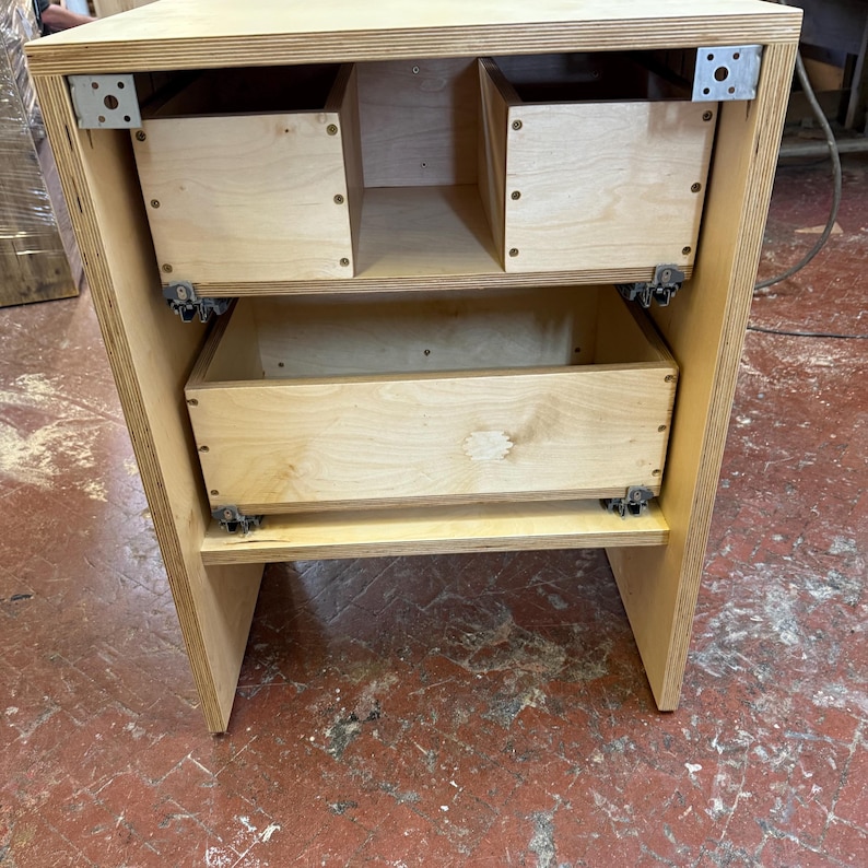 K&ouml;nnte beinhalten: Ein Holzschrank mit zwei Schubladen und zwei offenen F&auml;chern. Der Schrank ist aus hellem Holz gefertigt und hat Metallhalterungen zur Verst&auml;rkung. Die Schubladen sind rechteckig und haben ein schlichtes Design. Der Schrank dient wahrscheinlich zur Aufbewahrung.