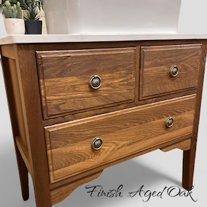 May include: A wooden cabinet with four drawers and ornate brass knobs. The cabinet has a light-coloured countertop and is labelled "Finish Aged Oak" in script. The wood grain is visible, and the cabinet has a warm, inviting tone.