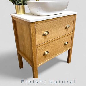 May include: A natural oak bathroom vanity featuring two drawers with brass-toned cup pulls, topped with a white rectangular countertop and a white ceramic vessel sink. A small green plant in a gold pot sits on the left side of the vanity top.
