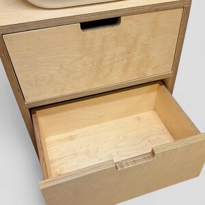 K&ouml;nnte beinhalten: Ein helles Holzschrank mit zwei Schubladen. Die obere Schublade ist geschlossen, w&auml;hrend die untere Schublade teilweise ge&ouml;ffnet ist und das Innere freigibt. Der Schrank hat ein schlichtes, modernes Design.