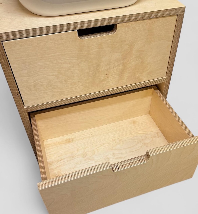 K&ouml;nnte beinhalten: Ein helles Holzschrank mit zwei Schubladen. Die obere Schublade ist geschlossen, w&auml;hrend die untere Schublade teilweise ge&ouml;ffnet ist und das Innere freigibt. Der Schrank hat ein schlichtes, modernes Design.