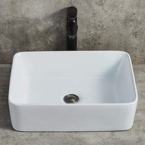 May include: White ceramic bathroom sink with a rectangular shape and a single drain. The sink has a smooth, glossy finish and is set against a grey background. A black faucet is visible above the sink.