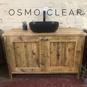 K&ouml;nnte beinhalten: Ein rustikaler Badezimmer-Waschtisch aus Holz mit zwei Schrankt&uuml;ren und einem schwarzen Keramikwaschbecken. Der Waschtisch hat eine gealterte Holzoptik und ist aus recyceltem Holz gefertigt. Die Schrankt&uuml;ren haben ein schlichtes Design mit einem einzigen Panel und einem Metallknopf.