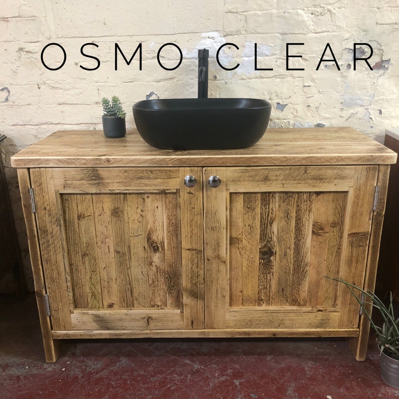K&ouml;nnte beinhalten: Ein rustikaler Badezimmer-Waschtisch aus Holz mit zwei Schrankt&uuml;ren und einem schwarzen Keramikwaschbecken. Der Waschtisch hat eine gealterte Holzoptik und ist aus recyceltem Holz gefertigt. Die Schrankt&uuml;ren haben ein schlichtes Design mit einem einzigen Panel und einem Metallknopf.