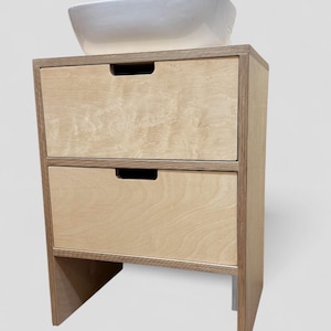 K&ouml;nnte beinhalten: Ein helles Holzwaschbeckenunterschrank mit zwei Schubladen und einem wei&szlig;en Keramikwaschbecken. Der Schrank hat eine rechteckige Form mit nat&uuml;rlicher Holzmaserung. Die Schubladen haben ein einfaches, eingelassenes Griffdesign.