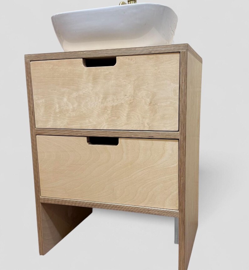 K&ouml;nnte beinhalten: Ein helles Holzwaschbeckenunterschrank mit zwei Schubladen und einem wei&szlig;en Keramikwaschbecken. Der Schrank hat eine rechteckige Form mit nat&uuml;rlicher Holzmaserung. Die Schubladen haben ein einfaches, eingelassenes Griffdesign.
