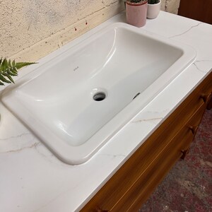 May include: White ceramic bathroom sink with a single drain hole, set into a white countertop with brown wood cabinets underneath. The sink has the word "VIVA" printed on the bottom.