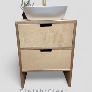 K&ouml;nnte beinhalten: Ein helles Holzwaschbeckenunterschrank mit zwei Schubladen und einem wei&szlig;en Keramikwaschbecken. Der Schrank hat eine transparente Oberfl&auml;che und ein minimalistisches Design. Oben steht eine kleine Pflanze. Der Text "Finish: Clear" steht unten.