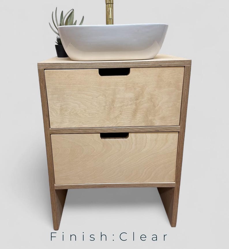 K&ouml;nnte beinhalten: Ein helles Holzwaschbeckenunterschrank mit zwei Schubladen und einem wei&szlig;en Keramikwaschbecken. Der Schrank hat eine transparente Oberfl&auml;che und ein minimalistisches Design. Oben steht eine kleine Pflanze. Der Text "Finish: Clear" steht unten.