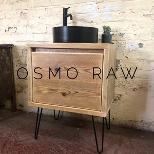 K&ouml;nnte beinhalten: Ein rustikaler Badezimmer-Waschtisch aus Holz mit einem schwarzen Keramikwaschbecken und einem Einhebelmischer. Der Waschtisch hat eine Schublade und steht auf Haarnadelbeinen. Der Text "OSMO RAW" ist auf der Vorderseite des Waschtisches sichtbar.