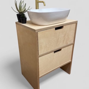 K&ouml;nnte beinhalten: Ein moderner Badezimmer-Waschtisch mit hellem Holzfinish. Er verf&uuml;gt &uuml;ber ein wei&szlig;es, rechteckiges Waschbecken, einen goldenen Wasserhahn und zwei Schubladen. Kleine Topfpflanzen stehen auf der Arbeitsplatte. Eine stilvolle Erg&auml;nzung f&uuml;r jedes Badezimmer.