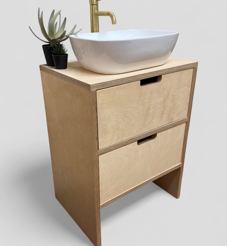 K&ouml;nnte beinhalten: Ein moderner Badezimmer-Waschtisch mit hellem Holzfinish. Er verf&uuml;gt &uuml;ber ein wei&szlig;es, rechteckiges Waschbecken, einen goldenen Wasserhahn und zwei Schubladen. Kleine Topfpflanzen stehen auf der Arbeitsplatte. Eine stilvolle Erg&auml;nzung f&uuml;r jedes Badezimmer.