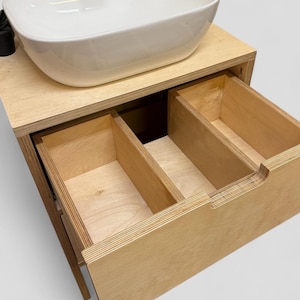 K&ouml;nnte beinhalten: Modernes Badezimmer-Waschbeckenunterschrank mit einem wei&szlig;en, oval geformten Waschbecken. Der Schrank ist aus hellem Holz gefertigt und verf&uuml;gt &uuml;ber zwei offene Schubladen. Das Design ist minimalistisch und funktional.