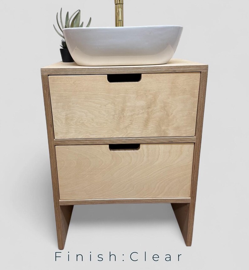 K&ouml;nnte beinhalten: Ein helles Holzwaschbeckenunterschrank mit zwei Schubladen und einem wei&szlig;en Keramikwaschbecken. Der Schrank hat eine transparente Oberfl&auml;che und ein minimalistisches Design. Oben steht eine kleine Pflanze. Der Text "Finish: Clear" steht unten.