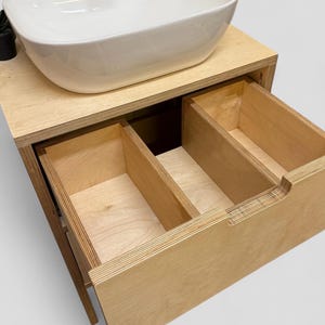 K&ouml;nnte beinhalten: Ein moderner Badezimmer-Waschtisch mit einem wei&szlig;en, oval geformten Waschbecken. Der Waschtisch ist aus hellem Holz gefertigt und verf&uuml;gt &uuml;ber zwei offene Schubladen. Das Design ist minimalistisch und funktional.