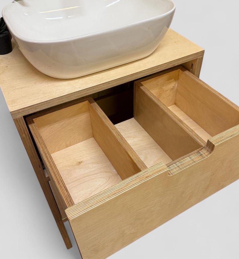 K&ouml;nnte beinhalten: Ein moderner Badezimmer-Waschtisch mit einem wei&szlig;en, oval geformten Waschbecken. Der Waschtisch ist aus hellem Holz gefertigt und verf&uuml;gt &uuml;ber zwei offene Schubladen. Das Design ist minimalistisch und funktional.