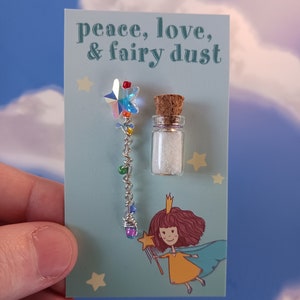 May include: A card with the text "peace, love, & fairy dust" printed on it. A small glass bottle filled with glitter and a silver wire wand with a rainbow star on top are attached to the card.
