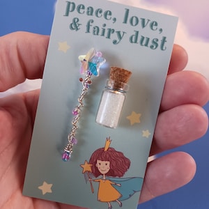May include: A blue card with the text "peace, love, & fairy dust" printed on it. The card features a small glass bottle filled with glitter and a silver wand with a iridescent star on top.