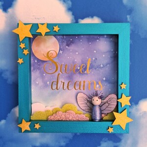 May include: A framed artwork depicting a night scene with a full moon, stars, and the gold text "Sweet dreams". A fairy figurine with purple wings and a blue dress is in the frame, with yellow star accents and clouds.
