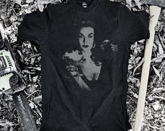 May include: Black graphic t-shirt with a grayscale image of a woman holding a glass, with a skull and a chainsaw and axe in the background. The shirt is on a bed of leaves.