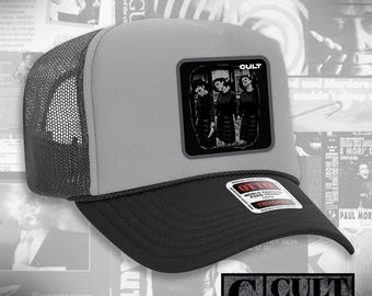 May include: A gray and black trucker hat with a black brim and mesh back. The hat features a square patch with the word "CULT" and a black and white image of three people. The hat also has a "CULT RATED" logo.